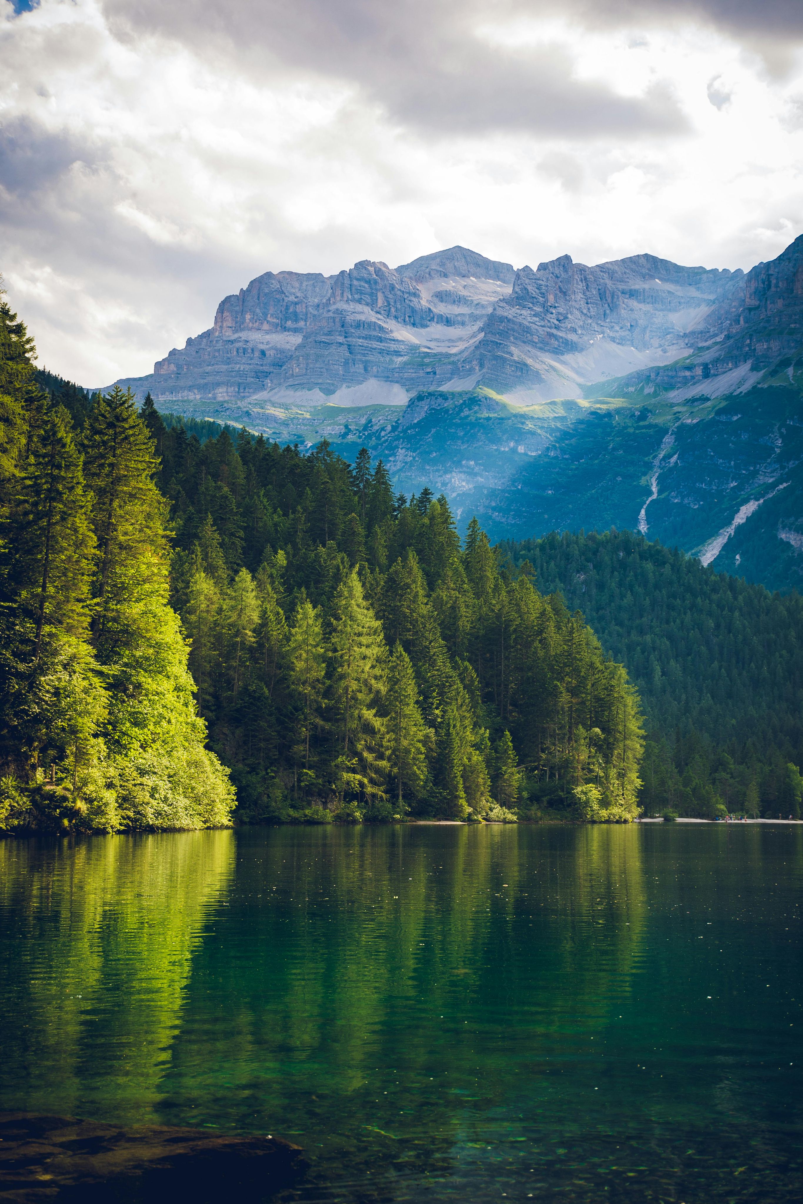 A serene mountain lake surrounded by dense evergreen forest with towering rocky peaks in the background under a cloudy sky.